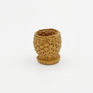 Puede incluir: Una pequeña cesta tejida de color marrón claro. La cesta tiene forma redondeada con la parte superior ensanchada y un patrón tejido texturizado. La base es ligeramente más ancha que el cuerpo.