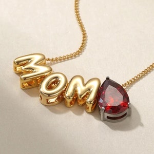 Peut inclure: Collier en or avec un pendentif en pierre rouge et le mot "MOM" écrit en lettres dorées.