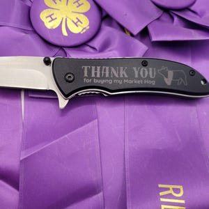 May include: A folding knife with a silver blade and a black handle. The handle is engraved with the words "THANK YOU for buying my Market Hog" and a pig illustration. The knife rests on a purple ribbon.