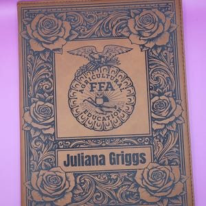May include: A brown leather notebook with a decorative floral border and the name "Juliana Griggs" engraved on the front. The FFA emblem is centered on the cover, with an eagle at the top.