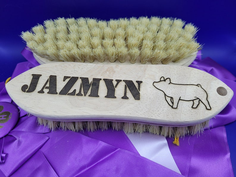 May include: A wooden brush with a pig silhouette and the name "Jazmyn" engraved on it. The brush has natural bristles and is designed for grooming animals.