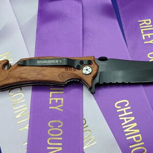 May include: A black folding knife with a brown wooden handle. The knife has a partial serrated blade and a safety lock. The knife is labeled "BISON RIVER" and "SCOTTA".