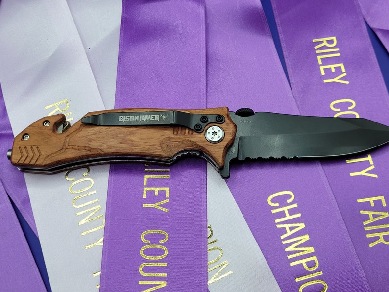 May include: A black folding knife with a brown wooden handle. The knife has a partial serrated blade and a safety lock. The knife is labeled "BISON RIVER" and "SCOTTA".