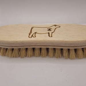 May include: A light-colored wooden grooming brush with a curved handle and natural bristles. The brush features a laser-etched outline of a cow on the top. The bristles are a light tan color.