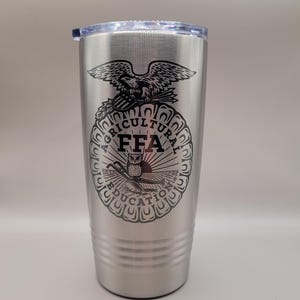 May include: A silver insulated tumbler with a black FFA emblem. The emblem features an eagle, owl, and the words "FFA AGRICULTURAL EDUCATION". The tumbler has a clear lid and is designed to hold approximately 20 ounces.