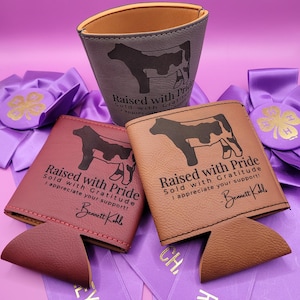 May include: A collection of drink holders in shades of brown and gray, featuring a cow silhouette and the text "Raised with Pride." The holders are displayed with purple ribbons and a 4-H emblem.