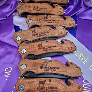 May include: A set of seven wooden handled pocket knives with engraved text and animal silhouettes. The knives are engraved with the text "Grand Champion" and the animal category, such as "Market Steer", "Commercial Ewe", "Commercial Doe", "Billy Goat", "Market Goat", "Market Lamb", and "Commercial Heifer".