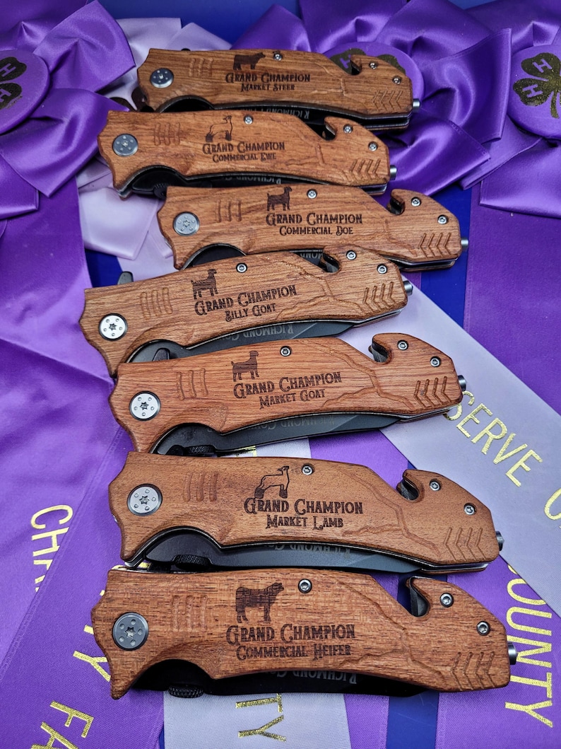 May include: A set of seven wooden handled pocket knives with engraved text and animal silhouettes. The knives are engraved with the text "Grand Champion" and the animal category, such as "Market Steer", "Commercial Ewe", "Commercial Doe", "Billy Goat", "Market Goat", "Market Lamb", and "Commercial Heifer".