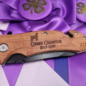 May include: A wooden handled pocket knife with a black blade. The handle is engraved with a silhouette of a goat and the text "Grand Champion Billy Goat".