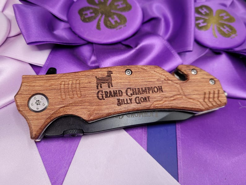 May include: A wooden handled pocket knife with a black blade. The handle is engraved with a silhouette of a goat and the text "Grand Champion Billy Goat".