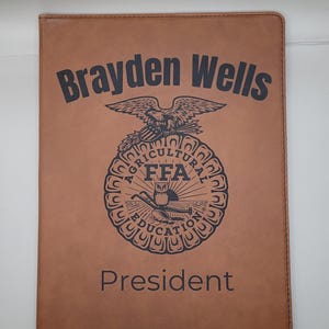 May include: A brown leatherette portfolio with the name "Brayden Wells" and the FFA emblem. The emblem features an eagle, an owl, and the words "Agricultural Education" and "FFA". The word "President" is printed below the emblem.