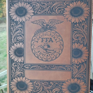 May include: A brown leather-bound notebook with a decorative black floral design and the FFA emblem. The cover features sunflowers in each corner and the words "AGRICULTURAL EDUCATION". The notebook has a blank rectangular space for personalization.