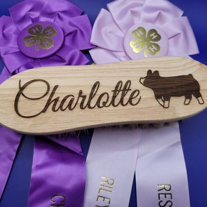 May include: A wooden pig grooming brush with the name "Charlotte" engraved on it. The brush is surrounded by two purple and white ribbons with gold lettering.