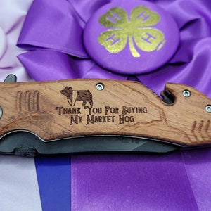 May include: A wooden pocket knife with a black blade and a  "Thank You for Buying My Market Hog" inscription. The knife has a small pig silhouette and a bottle opener.