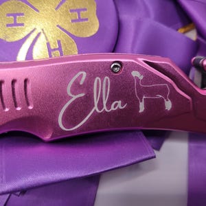 May include: A pink multi-tool with the name "Ella" in script and a sheep silhouette. The tool is set against a backdrop of purple ribbons and a purple and gold emblem. The tool has a blade and other features.