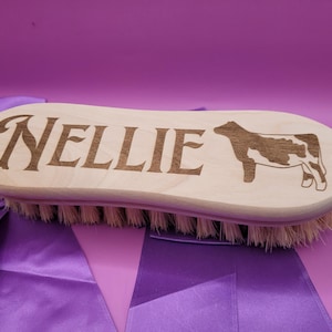 May include: A light-colored wooden grooming brush featuring the engraved name "NELLIE" in a decorative script font alongside a laser-etched silhouette of a cow. The brush has stiff, light-brown bristles and a small circular hole at one end.