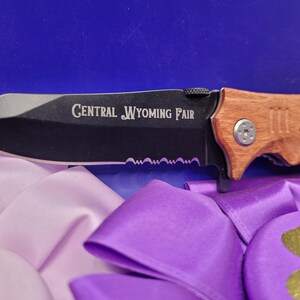 May include: A black folding knife with a wooden handle. The blade has a serrated edge and the handle is engraved with "Central Wyoming Fair" and "Thank you for my market".