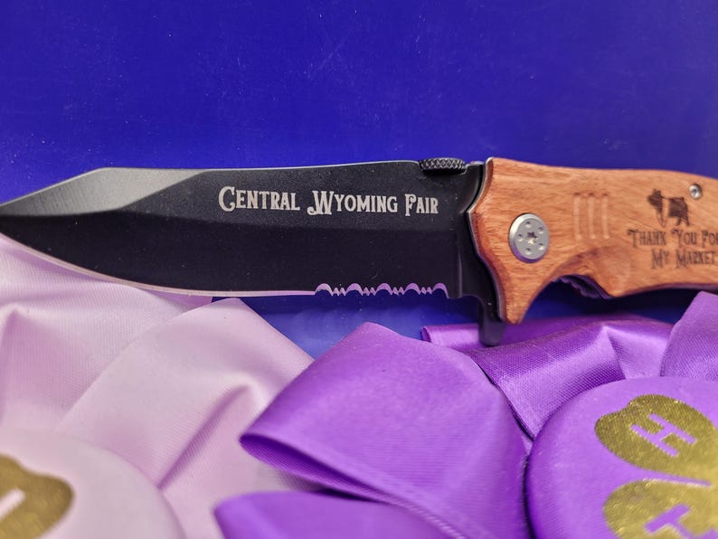 May include: A black folding knife with a wooden handle. The blade has a serrated edge and the handle is engraved with "Central Wyoming Fair" and "Thank you for my market".