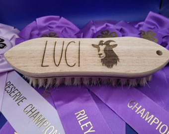 May include: A wooden grooming brush with a white bristle brush. The brush is engraved with the name "LUCI" and a goat silhouette. The brush is lying on top of purple ribbons with gold lettering.