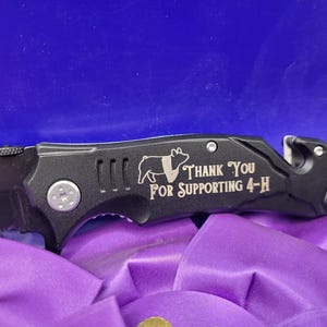 May include: Black folding knife with a silver engraved pig and the text "Thank You For Supporting 4-H".