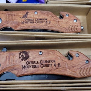 May include: Two wooden pocket knives with black blades. The handles are engraved with the text "Overall Champion Mountrail County 4-H".