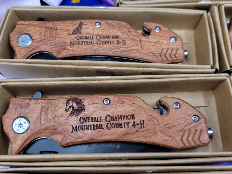 May include: Two wooden pocket knives with black blades. The handles are engraved with the text "Overall Champion Mountrail County 4-H".