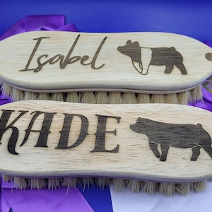 May include: Two wooden pig brushes with black bristles. The top brush has the name "Isabel" and a silhouette of a pig. The bottom brush has the name "KADE" and a silhouette of a pig.