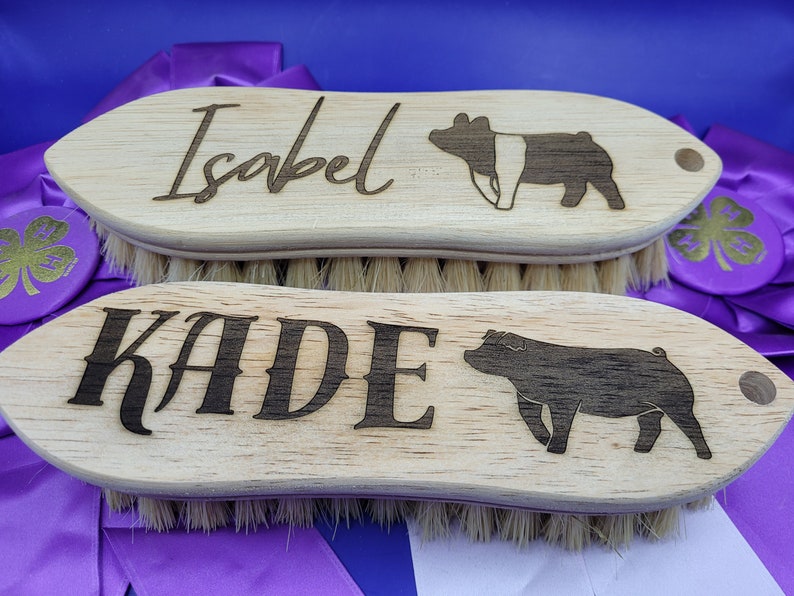 May include: Two wooden pig brushes with black bristles. The top brush has the name "Isabel" and a silhouette of a pig. The bottom brush has the name "KADE" and a silhouette of a pig.