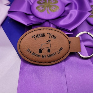 May include: Brown leather keychain with a silver key ring. The keychain has an oval-shaped tag with the text "Thank You For Buying My Market Lamb" and a silhouette of a lamb.
