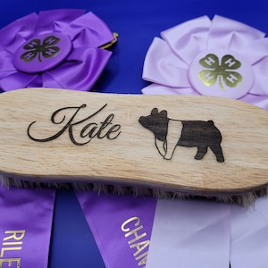 May include: A wooden pig show brush with the name "Kate" engraved on it. The brush is surrounded by purple and white ribbons with gold lettering that says "Champion" and "Reserve Champion".