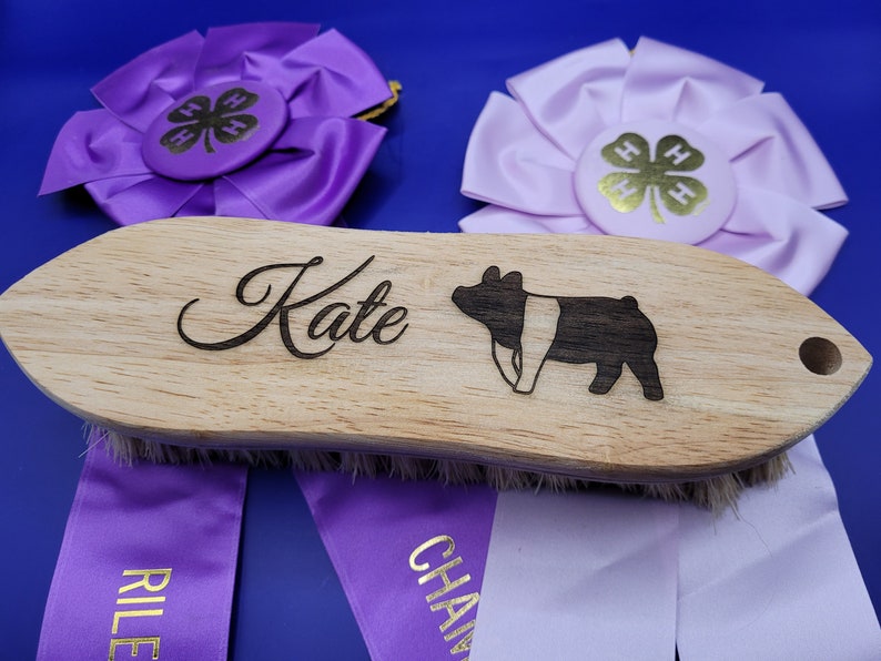 May include: A wooden pig show brush with the name "Kate" engraved on it. The brush is surrounded by purple and white ribbons with gold lettering that says "Champion" and "Reserve Champion".