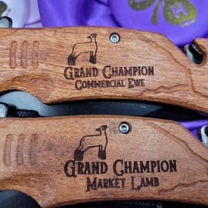 May include: Two wooden pocket knives with engraved sheep and text. The top knife reads "Grand Champion Commercial Ewe". The bottom knife reads "Grand Champion Market Lamb".