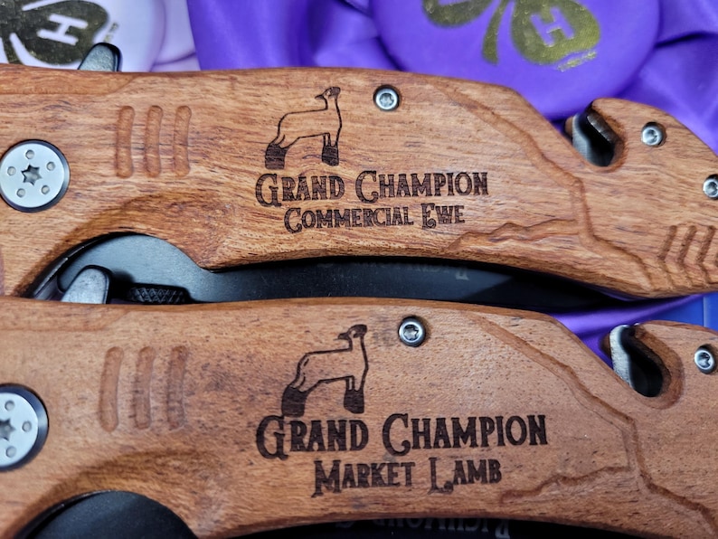 May include: Two wooden pocket knives with engraved sheep and text. The top knife reads "Grand Champion Commercial Ewe". The bottom knife reads "Grand Champion Market Lamb".