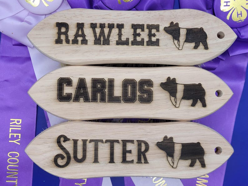 May include: Three wooden plaques with the names "Rawlee", "Carlos", and "Sutter" engraved on them. Each plaque features a black and white pig illustration.