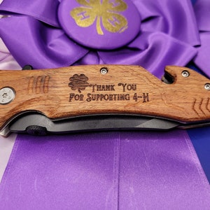 May include: A wooden pocket knife with a black blade and a wood handle. The handle is engraved with a four-leaf clover, the words "Thank You For Supporting 4-H", and a small silver screw.