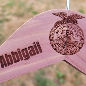 May include: A wooden hanger with the name "Abbigail" engraved in a dark brown font. The hanger features a detailed FFA emblem with an eagle and the words "AGRICULTURE EDUCATION" in a circular design.
