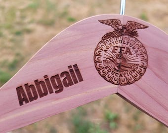 May include: A wooden hanger with the name "Abbigail" engraved in a dark brown font. The hanger features a detailed FFA emblem with an eagle and the words "AGRICULTURE EDUCATION" in a circular design.