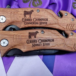 May include: Two wooden handled pocket knives with black blades. The handles are engraved with the text "Grand Champion Commercial Heifer" and "Grand Champion Market Steer".