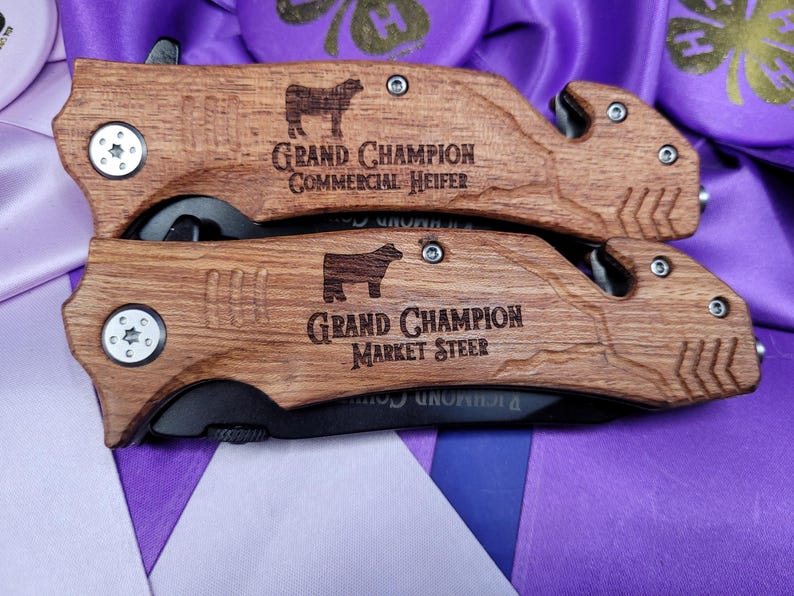 May include: Two wooden handled pocket knives with black blades. The handles are engraved with the text "Grand Champion Commercial Heifer" and "Grand Champion Market Steer".