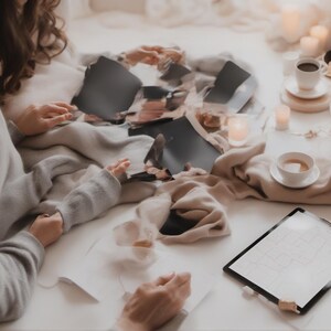May include: A gray sweater and beige fabric draped over a surface with scattered dark paper fragments, two white ceramic cups on saucers, small lit candles, and a black clipboard holding a white sheet of paper with a faint grid pattern.