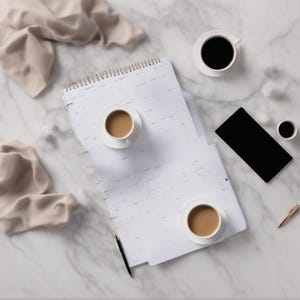 Puede incluir: Una agenda blanca con encuadernaci&oacute;n en espiral y dise&ntilde;o de cuadr&iacute;cula, dos tazas de caf&eacute; con leche sobre platillos, una taza de caf&eacute; negro sobre platillo, una taza negra peque&ntilde;a, un tel&eacute;fono inteligente negro, un bol&iacute;grafo dorado y una tela beige.
