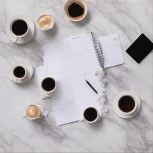 May include: A spiral-bound white paper planner with a black pen resting on the page, surrounded by seven white ceramic cups filled with black coffee or latte, and a small black rectangular object, all arranged on a white marble surface.