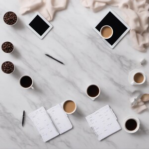 Puede incluir: Una superficie de mármol blanco dispuesta con tres cuencos pequeños de granos de café tostado oscuro, dos tabletas, dos agendas abiertas, un bolígrafo negro y cinco tazas de cerámica blanca con café negro o con leche, junto a tela clara.