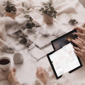 May include: Two black tablets with illuminated screens displaying digital maps and interface elements, positioned alongside a white ceramic cup filled with dark liquid and two small potted green plants on a soft, textured cream-coloured fabric.
