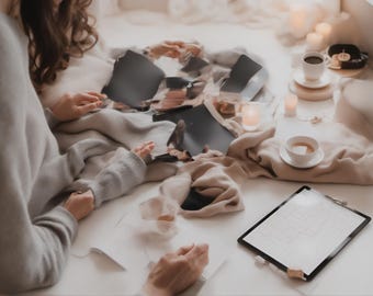 May include: A gray sweater and beige fabric draped over a surface with scattered dark paper fragments, two white ceramic cups on saucers, small lit candles, and a black clipboard holding a white sheet of paper with a faint grid pattern.