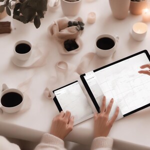 K&ouml;nnte beinhalten: Ein gro&szlig;es Tablet und ein kleineres Tablet mit einer Raster-Kalenderansicht auf einer wei&szlig;en Oberfl&auml;che, begleitet von drei wei&szlig;en Keramiktassen mit schwarzem Kaffee, kleinen Schalen mit dunklen Beeren und mehreren brennenden Kerzen.