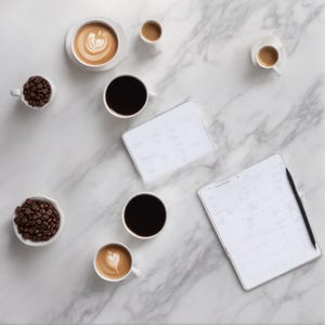 K&ouml;nnte beinhalten: Eine Sammlung von Kaffeeartikeln, darunter zwei wei&szlig;e Schalen mit dunkel ger&ouml;steten Kaffeebohnen, zwei Tassen schwarzer Kaffee, zwei Lattes mit Milchschaumkunst und zwei kleine Espressotassen. Zwei wei&szlig;e Planer mit schwarzen Stiften liegen daneben.