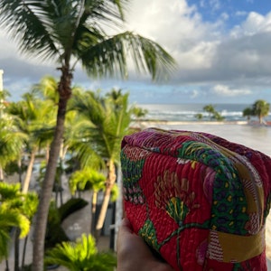 May include: A red quilted makeup bag with a floral pattern. The bag has a zipper closure and a yellow trim. The bag is being held in a hand against a background of palm trees and a blue sky.