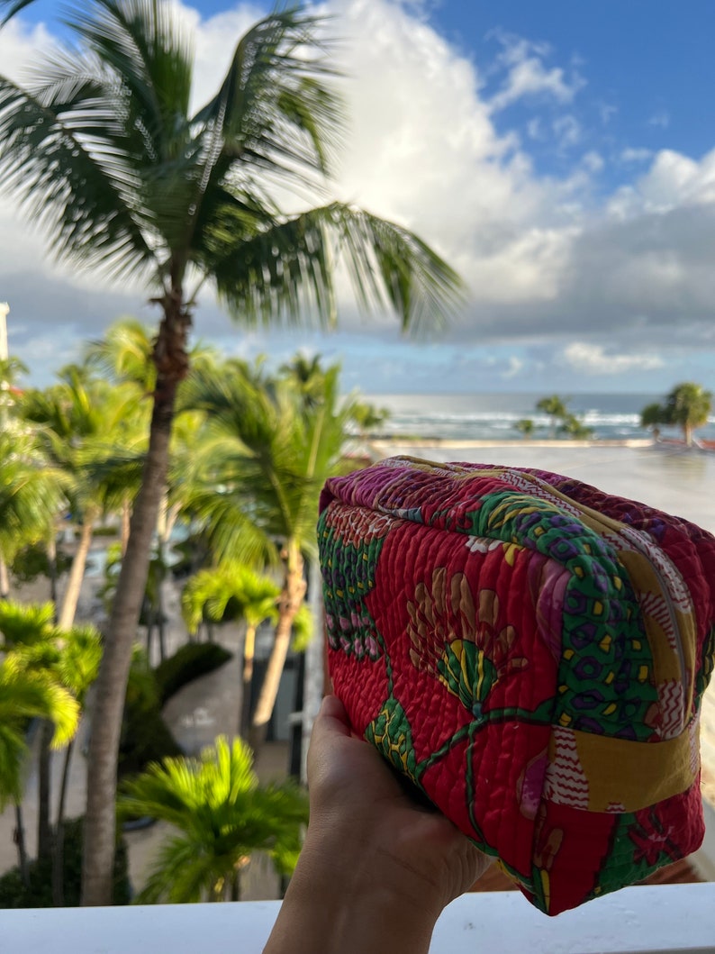 May include: A red quilted makeup bag with a floral pattern. The bag has a zipper closure and a yellow trim. The bag is being held in a hand against a background of palm trees and a blue sky.