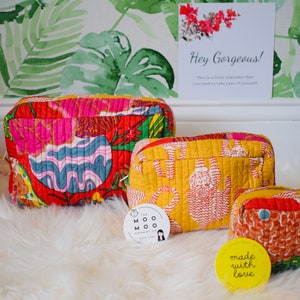 May include: Three quilted cosmetic bags with vibrant floral and abstract patterns in red, yellow, and blue. The bags are on a fluffy white surface, with a sign that says "Hey Gorgeous!" in the background.